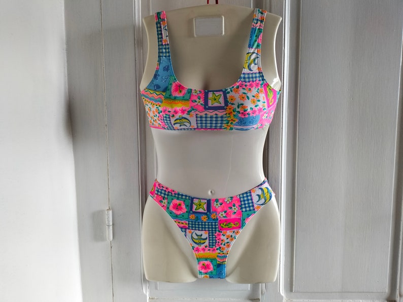 Puede incluir: Un traje de ba&ntilde;o de dos piezas con un dise&ntilde;o de patchwork. La parte superior es de estilo cuello redondo con tirantes anchos. La parte inferior es de corte alto. El traje de ba&ntilde;o presenta una mezcla de estampados florales, cuadros y geom&eacute;tricos en rosa, azul, amarillo y verde.
