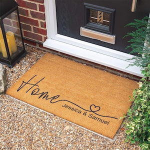 Puede incluir: Una alfombra de coco marrón con la palabra "Home" escrita en cursiva. Debajo de la palabra "Home" hay un corazón decorativo y el texto "Jessica & Samuel".