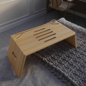 May include: A light wood laptop stand with cutouts for ventilation and a handle on the side. The stand is on a grey fluffy blanket.