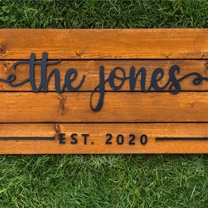 May include: A wooden sign with the text "the jones est. 2020" in black lettering. The sign is made of multiple wooden planks and has a rustic, farmhouse style.