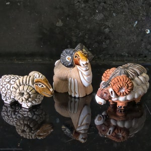 Puede incluir: Tres figuritas de ovejas de cerámica con detalles intrincados. Las ovejas son marrones, blancas y negras con cuernos rizados. Las ovejas están de pie sobre una superficie negra.