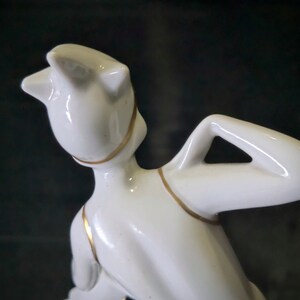 May include: White porcelain figurine of a woman with a gold trim around her neck and arms. The woman is posed with her arm raised above her head.