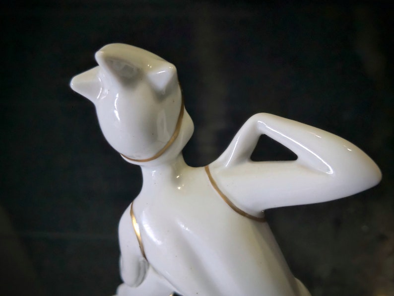 May include: White porcelain figurine of a woman with a gold trim around her neck and arms. The woman is posed with her arm raised above her head.