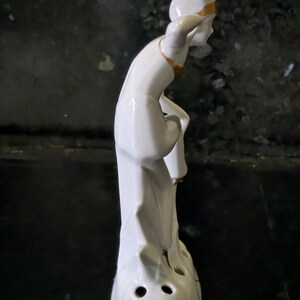 May include: White porcelain figurine of a person with a pointed hat and gold accents. The figurine is standing on a base with three holes.