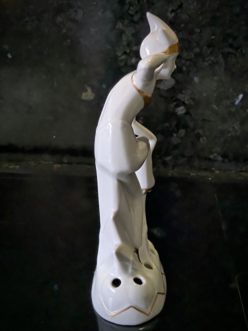 May include: White porcelain figurine of a person with a pointed hat and gold accents. The figurine is standing on a base with three holes.