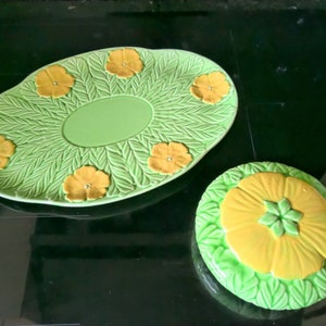 Puede incluir: Un plato de servir de cerámica verde con un diseño floral en relieve. El plato tiene un pozo central y está decorado con flores amarillas. También se muestra un plato más pequeño a juego con una sola flor amarilla.