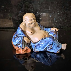 May include: A ceramic figurine of a smiling Buddha sitting cross-legged, wearing a blue and brown robe. The figurine is sitting on a brown cushion.