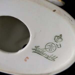May include: Close-up of the bottom of a white porcelain figurine. The mark reads "P.G. L&uuml;ntherfeld" with a crown and a sword.