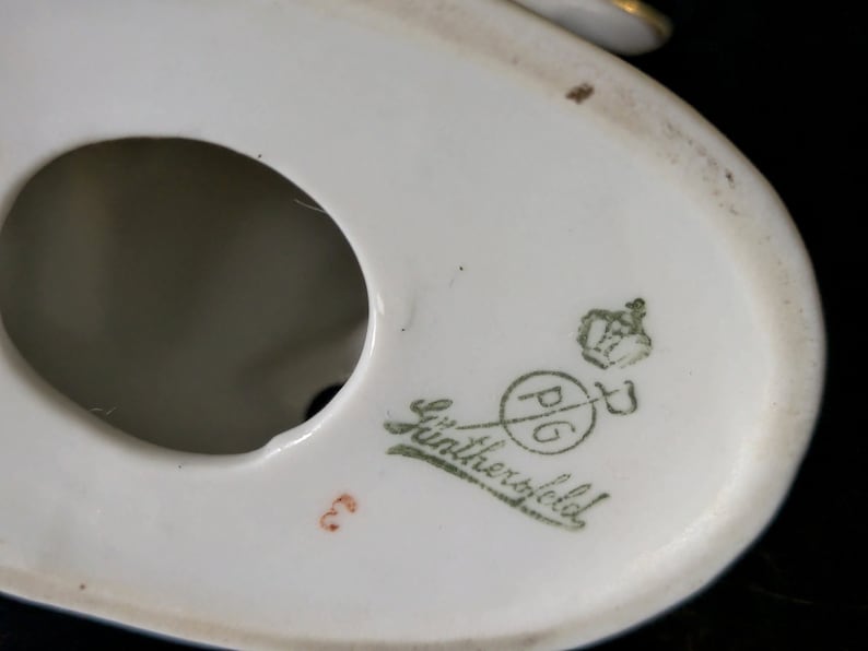 May include: Close-up of the bottom of a white porcelain figurine. The mark reads "P.G. L&uuml;ntherfeld" with a crown and a sword.