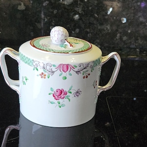 Könnte beinhalten: Weiße Porzellan-Zuckerdose mit Deckel. Die Schale hat ein grünes und rotes Blumenmuster mit einer rosa Rose in der Mitte. Der Deckel hat ein grünes und rotes Blumenmuster mit einem kleinen, runden Knopf.