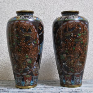 May include: Two antique cloisonné vases. The vases have a dark brown background with intricate floral and bird designs in various colors. The rims and bases are trimmed with gold-colored metal.