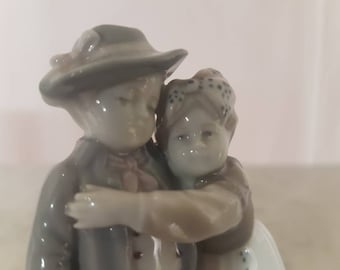 May include: A porcelain figurine of a couple embracing. The man is wearing a hat and a jacket, while the woman is wearing a dress with a floral pattern. The figurine is painted in shades of white, gray, and brown.