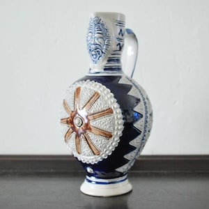 Peut inclure: Un pichet en céramique bleu et blanc avec un motif de roue marron. Le pichet a une anse et un bec.