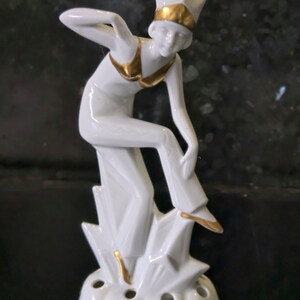 May include: White porcelain figurine of a woman in a gold-trimmed outfit, standing on a base with multiple holes. The figurine is in a stylized art deco style.