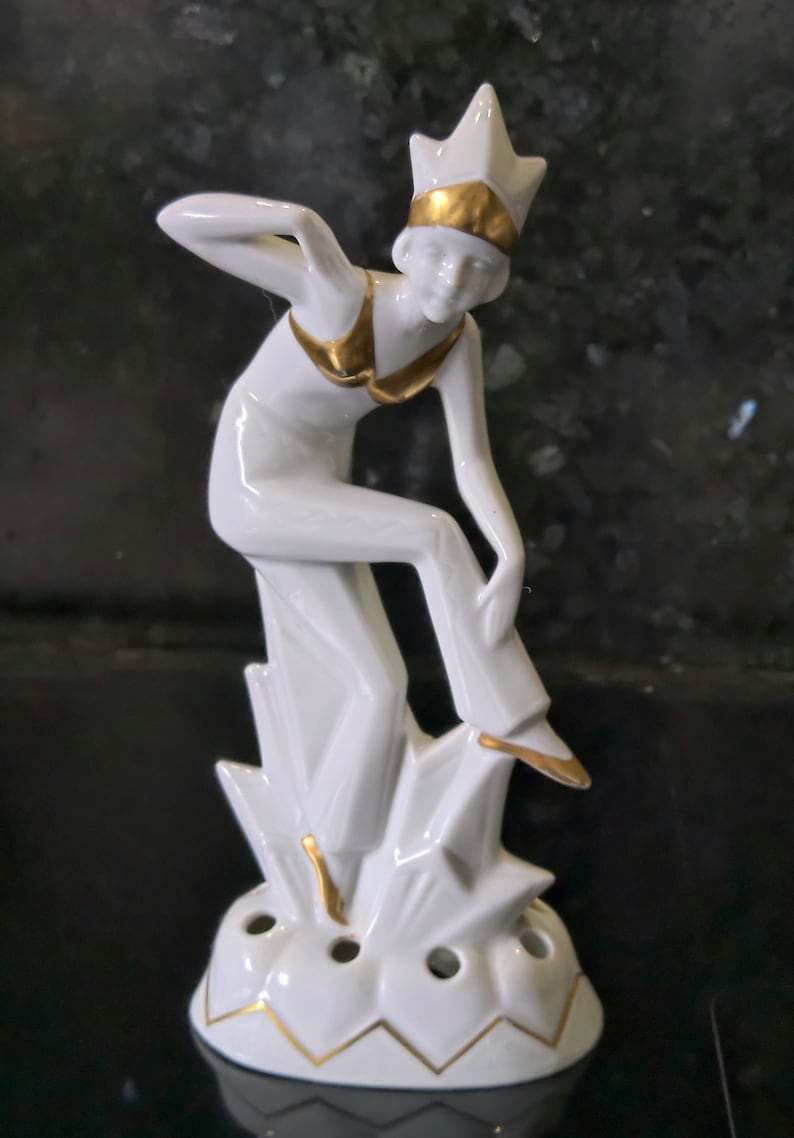 May include: White porcelain figurine of a woman in a gold-trimmed outfit, standing on a base with multiple holes. The figurine is in a stylized art deco style.