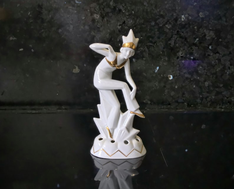 May include: White porcelain figurine of a woman in a gold crown, with gold accents, standing on a base with a gold trim.