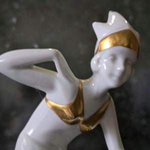 May include: A white porcelain figurine of a woman wearing a gold and white swimsuit and a gold headband. The figurine is in a dynamic pose, with one arm raised above her head.