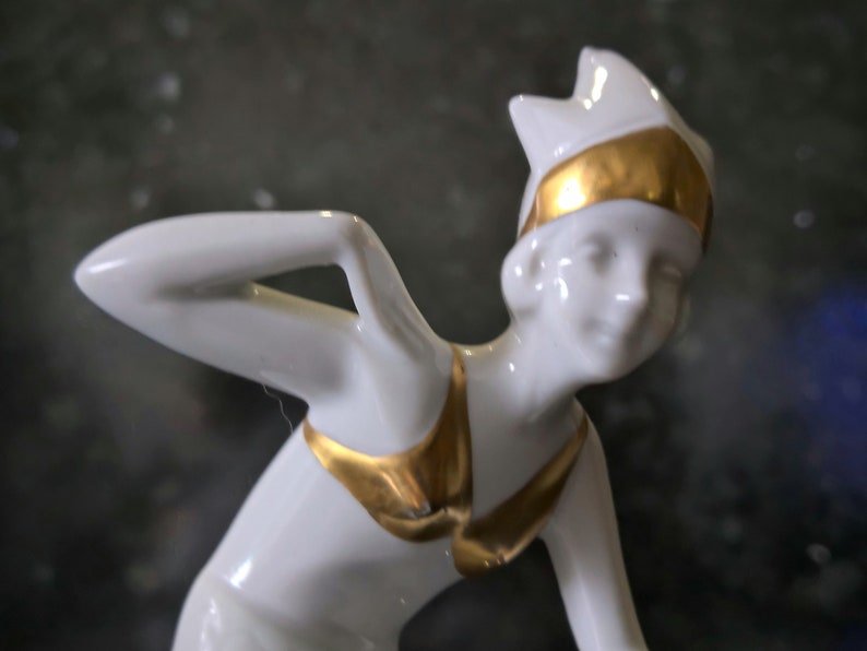 May include: A white porcelain figurine of a woman wearing a gold and white swimsuit and a gold headband. The figurine is in a dynamic pose, with one arm raised above her head.