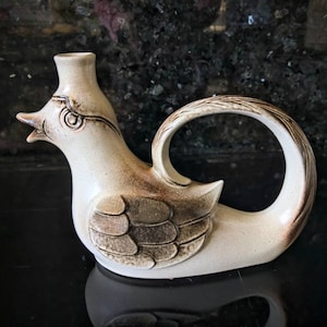 May include: A ceramic bird-shaped pitcher with a brown and white glaze. The pitcher has a spout and a handle that forms a loop around the back of the bird's body.