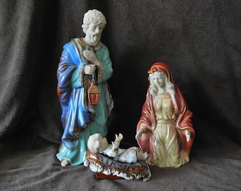 May include: A ceramic nativity scene figurine set featuring a blue robed Joseph, a red robed Mary, and a baby Jesus lying in a manger.