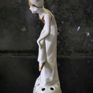 May include: White porcelain figurine of a woman wearing a hat with gold accents. The woman is holding a decorative object in her hand. The figurine has a base with four holes and gold accents.