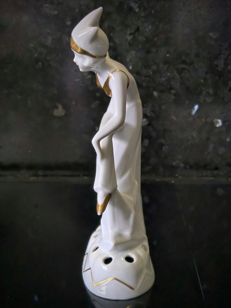 May include: White porcelain figurine of a woman wearing a hat with gold accents. The woman is holding a decorative object in her hand. The figurine has a base with four holes and gold accents.