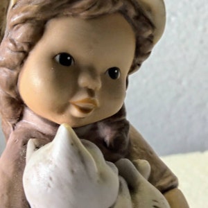 May include: A porcelain figurine of a young girl wearing a brown dress and a yellow hat, holding a white porcelain kitten.
