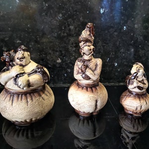Könnte beinhalten: Drei Keramikfiguren von Menschen mit übertriebenen Gesichtszügen. Die Figuren sind in Braun- und Beigetönen bemalt und haben ein rustikales Aussehen. Die größte Figur hält eine kleinere Figur in ihren Armen.