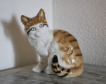 May include: A ceramic figurine of a brown and white tabby cat sitting with its tail curled around its legs. The cat has black eyes and a white face.