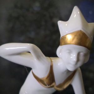 May include: White porcelain figurine of a woman wearing a gold crown and a gold swimsuit. The figurine is in a crouched position, with one arm raised above her head.