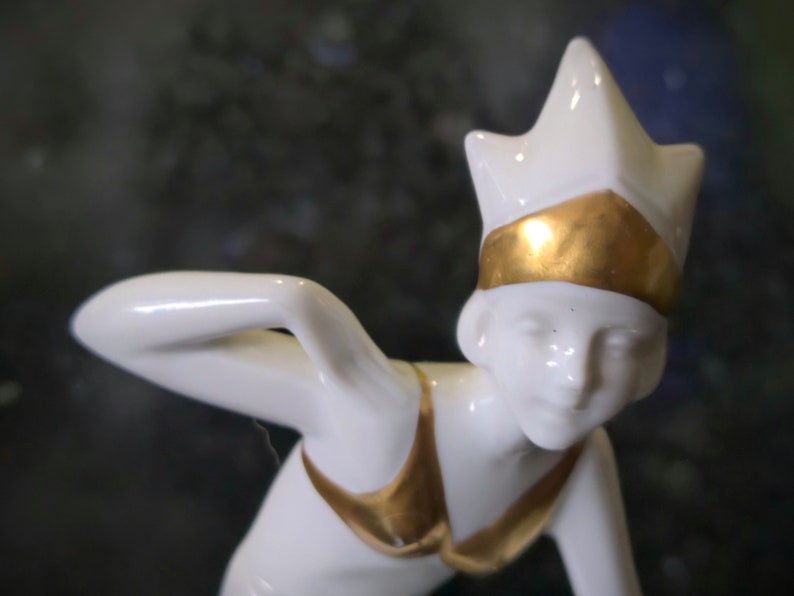 May include: White porcelain figurine of a woman wearing a gold crown and a gold swimsuit. The figurine is in a crouched position, with one arm raised above her head.
