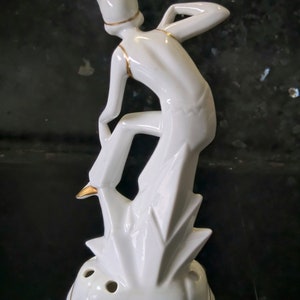 May include: White ceramic figurine of a stylized jester with gold accents standing on a geometric base with holes. The jester is wearing a pointed hat and is in a playful pose.