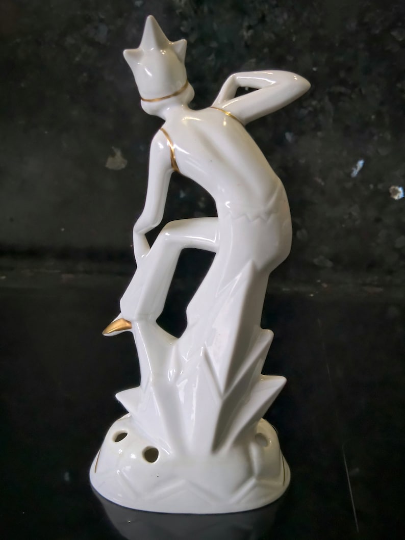 May include: White ceramic figurine of a stylized jester with gold accents standing on a geometric base with holes. The jester is wearing a pointed hat and is in a playful pose.