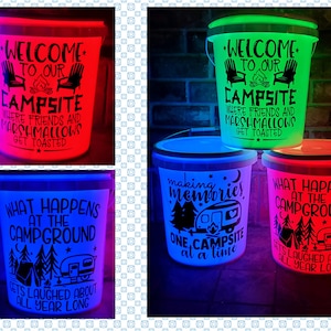 May include: Four white plastic buckets with different camping-themed designs and glowing LED lights in red, green, blue, and pink. The text on the buckets reads: "Welcome to our campsite where friends and marshmallows get toasted", "Making memories, one campsite at a time", "What happens at the campground gets laughed about all year long", and "Welcome to our campsite where friends and marshmallows get toasted".