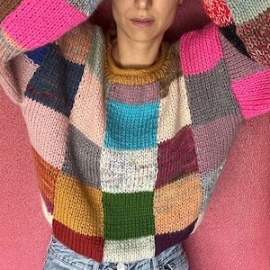 May include: A colourful patchwork sweater with a crew neck and long sleeves. The sweater features squares of pink, blue, yellow, and green. The person is wearing blue jeans.