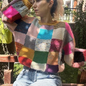 May include: A patchwork sweater in a variety of colors, including pink, blue, yellow, and green. The sweater has a crew neck and long sleeves. The person is wearing blue jeans and sitting on a wooden bench in a garden setting.