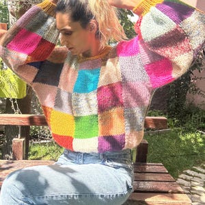 May include: A colorful patchwork sweater with a crew neck and long sleeves. The sweater features squares of pink, blue, yellow, green, and purple. The cuffs and neckline are a solid yellow color. The person is wearing blue jeans and sitting on a wooden bench.