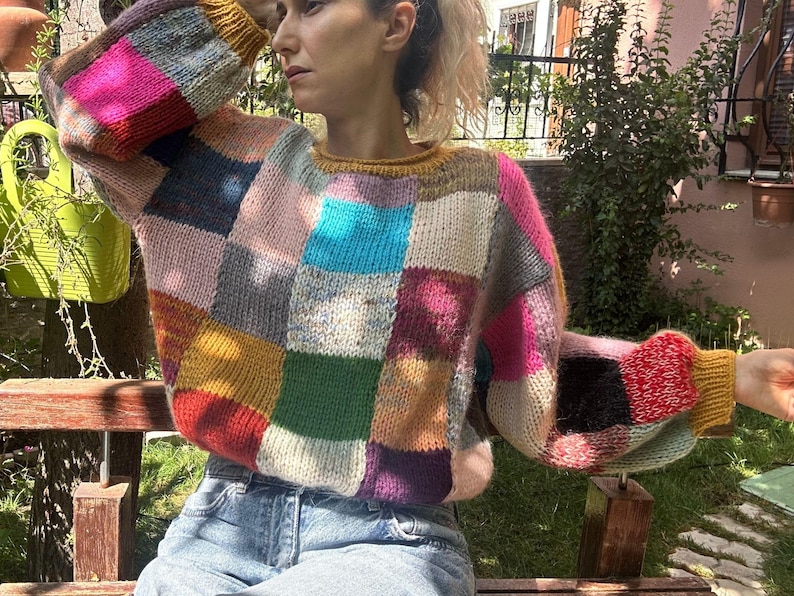 May include: A patchwork sweater in a variety of colors, including pink, blue, yellow, and green. The sweater has a crew neck and long sleeves. The person is wearing blue jeans and sitting on a wooden bench in a garden setting.