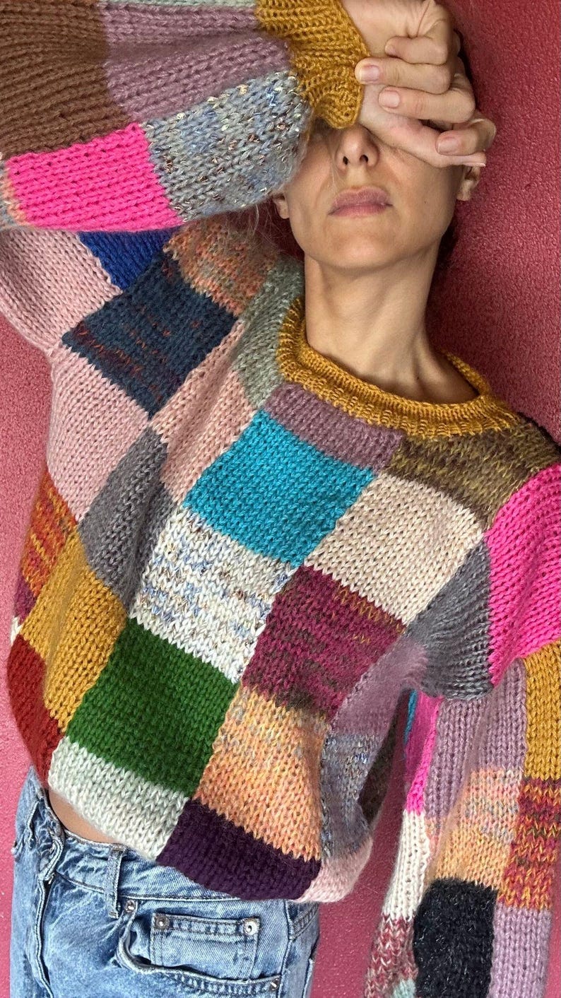 May include: A patchwork sweater in a variety of colors including pink, blue, green, and yellow. The sweater has a crew neck and long sleeves. The design is made of squares of different colored yarn.