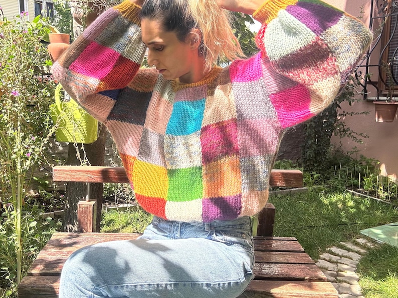 May include: A colorful patchwork sweater with a crew neck and long sleeves. The sweater features squares of pink, blue, yellow, green, and purple. The cuffs and neckline are a solid yellow color. The person is wearing blue jeans and sitting on a wooden bench.