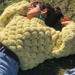 Pu&ograve; includere: Un maglione di lana giallo con un motivo a treccia spessa. Il maglione &egrave; oversize e ha una vestibilit&agrave; rilassata. La modella &egrave; sdraiata sull'erba indossando il maglione.