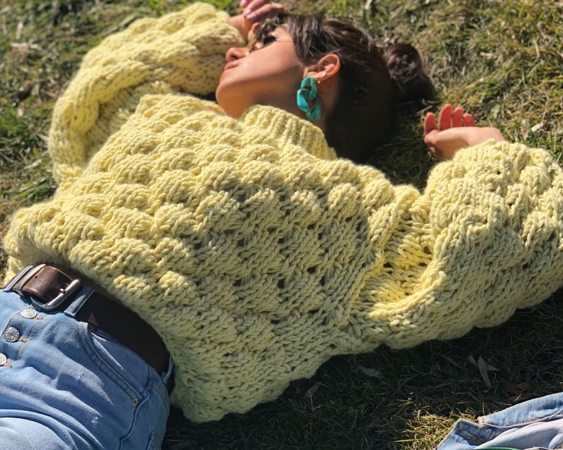 Pu&ograve; includere: Un maglione di lana giallo con un motivo a treccia spessa. Il maglione &egrave; oversize e ha una vestibilit&agrave; rilassata. La modella &egrave; sdraiata sull'erba indossando il maglione.