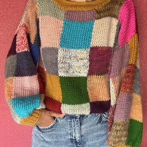 May include: A patchwork sweater in a variety of colors, including pink, blue, green, and yellow. The sweater has a round neckline and long sleeves. The person is wearing blue jeans.