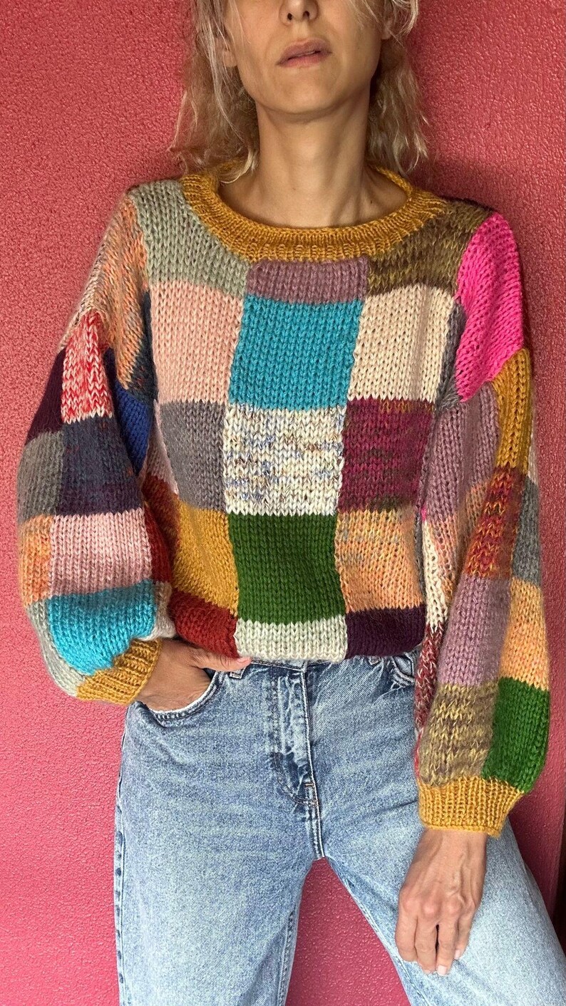 May include: A patchwork sweater in a variety of colors, including pink, blue, green, and yellow. The sweater has a round neckline and long sleeves. The person is wearing blue jeans.