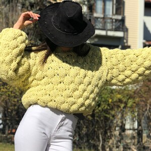 Pu&ograve; includere: Un maglione di lana giallo con un design testurizzato e gonfio. Il maglione ha un collo rotondo e maniche lunghe. La modella indossa un cappello nero.