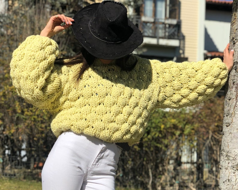 Pu&ograve; includere: Un maglione di lana giallo con un design testurizzato e gonfio. Il maglione ha un collo rotondo e maniche lunghe. La modella indossa un cappello nero.