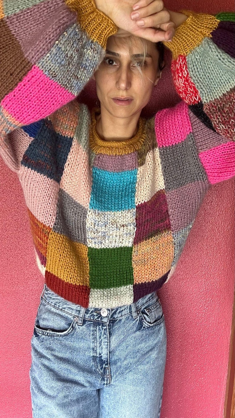 May include: A patchwork sweater in a variety of colors, including pink, blue, yellow, and green. The sweater has a crew neck and long sleeves. The person is wearing blue jeans.