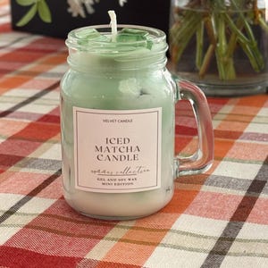 May include: A clear glass jar candle with a handle, filled with pale green wax. The label reads "Iced Matcha Candle" from the "Summer Collection". The candle is a mini edition, made with gel and soy wax.