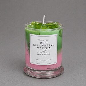 May include: A soy wax candle in a clear glass jar featuring a layered pink and green design topped with green wax ice cubes. The white label reads VELVET CANDLE ICED STRAWBERRY MATCHA latte SOY WAX CANDLE 180 g | 40 hours | 8 oz with a central wick.