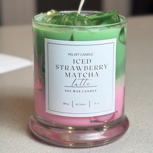 May include: A soy wax candle in a clear glass jar with a white label reading VELVET CANDLE ICED STRAWBERRY MATCHA latte SOY WAX CANDLE 180 g | 40 hours | 8 oz. The candle features layers of pink and green wax with green wax ice cubes.